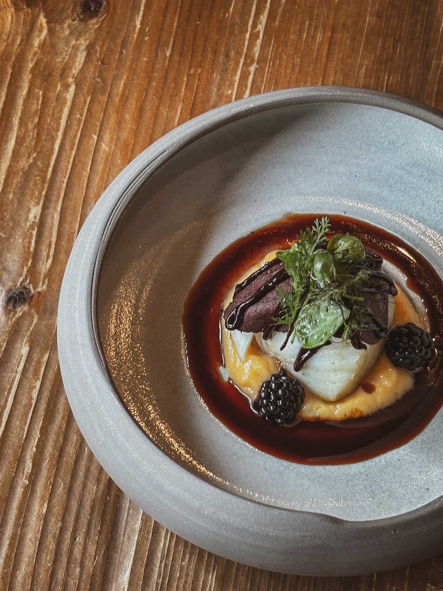 Angerichtetes Gourmetgericht in grauer Schale mit hellem Hauptbestandteil auf Püree, dunkler Sauce, Brombeeren und frischen Kräutern auf Holztisch.